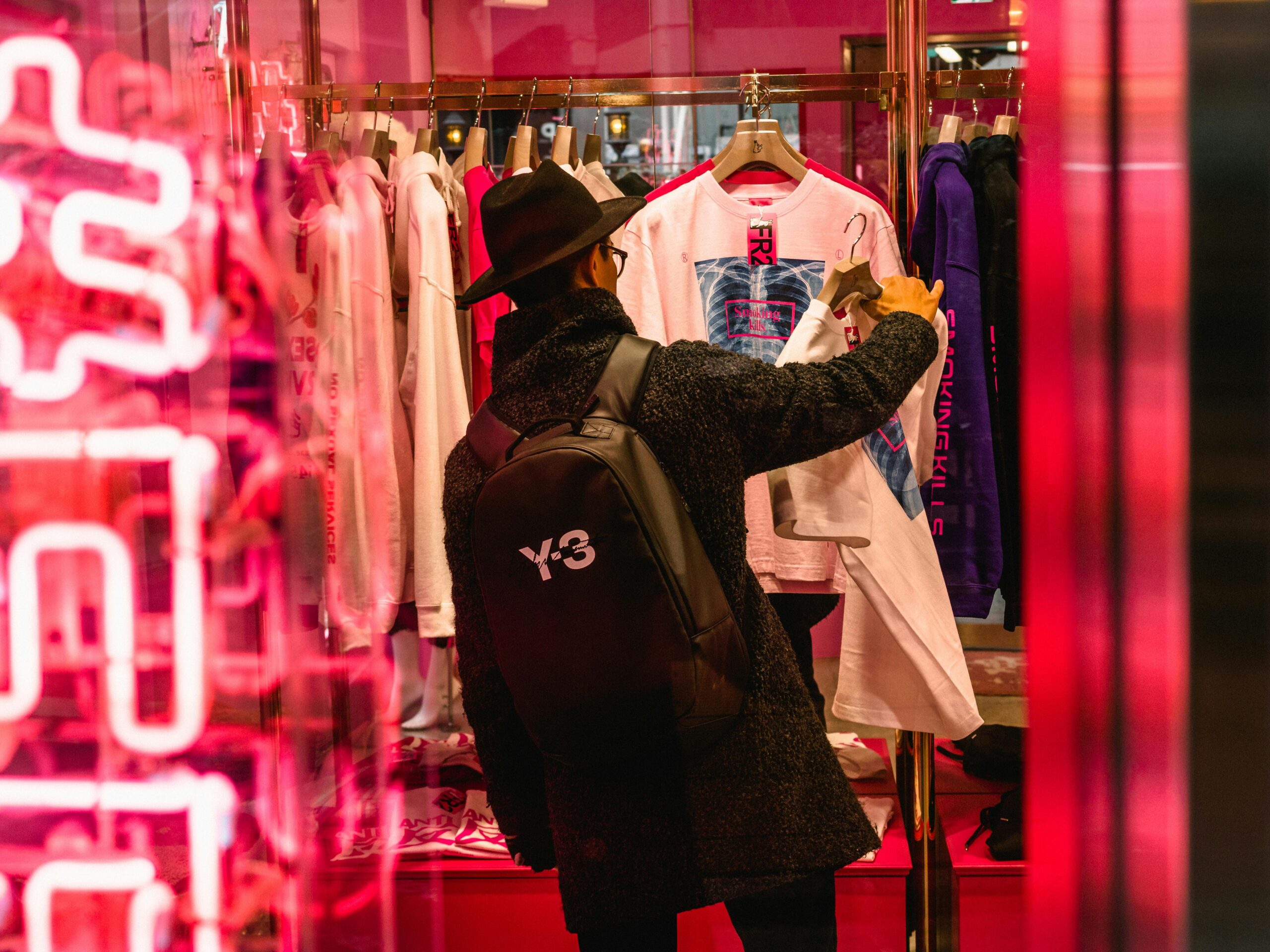A man in a black coat and hat shops for trendy clothing in a brightly lit boutique.